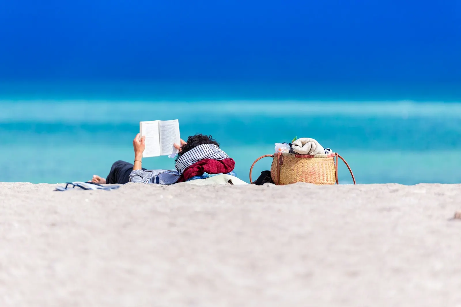 Eine Person liegt lesend am Strand, dahinter türkisfarbenes Meer