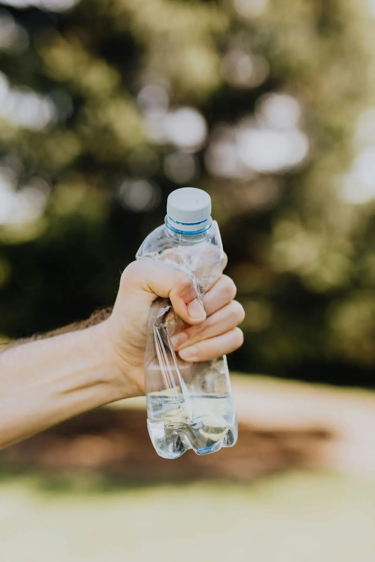 Crop unrecognizable man crumpling plastic bottle of water in hand against blurred background of green park trees on sunny summer day